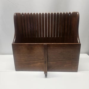 Puede incluir: Un revistero de madera marrón oscuro con respaldo de listones y base rectangular. El revistero tiene un asa en la parte trasera y una pequeña pieza de soporte en la parte delantera. La veta de la madera es visible.