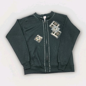 May include: Dark green zip-up sweatshirt with a front pocket and embroidered details. The sweatshirt features three plaid patches with the Green Bay Packers logo. The zipper is silver, and the collar and zipper line have decorative stitching.