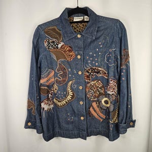 May include: A denim jacket with intricate, colorful embroidered and appliqued designs. The jacket features brown, tan, and animal print patterns, with gold-colored buttons and a leopard print collar lining. The jacket is hanging on a black hanger.