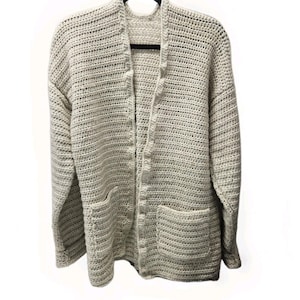 May include: A light beige, open-front crochet cardigan with two front pockets. The long-sleeved sweater is made of a thick, textured yarn and hangs on a black hanger against a white background. The cardigan has a relaxed fit.