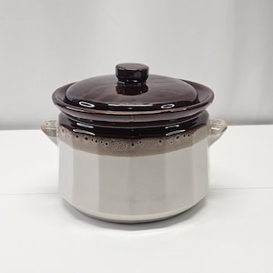 May include: A ceramic casserole dish with a dark brown lid and a white body. The dish has two small handles and a brown rim. The lid has a small knob on top. The dish is designed for cooking and serving.
