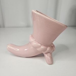 May include: A pink ceramic boot-shaped vase. The vase has a high heel and a decorative rope-like detail around the ankle. The opening of the vase is angled. The boot is a solid, soft pink colour.