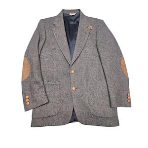 Puede incluir: Un blazer de tweed gris con coderas de ante marrón y botones de madera. El blazer tiene solapas con muescas, dos bolsillos delanteros y un forro azul oscuro. Un estilo clásico y vintage.