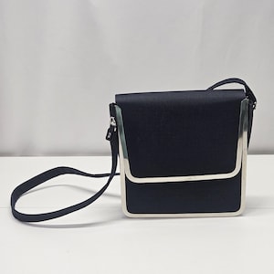 Puede incluir: Un bolso bandolera azul marino con forma rectangular y un marco de metal plateado. El bolso tiene una correa larga y fina y un cierre de solapa. El bolso está hecho de una tela texturizada.