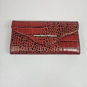 May include: A red, faux-crocodile skin wallet with a silver-colored bamboo-style clasp. The wallet has a flap closure and a textured surface. The wallet is rectangular in shape.