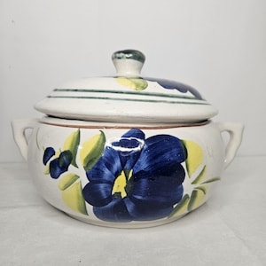 Puede incluir: Una sopera de cerámica blanca con tapa, con diseños florales azules y amarillos pintados a mano. La sopera tiene dos asas pequeñas y un pomo verde en la tapa. El diseño floral incluye una gran flor azul.