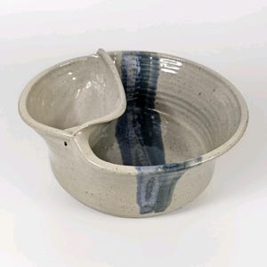 May include: A speckled ceramic bowl with a built-in side compartment. The bowl has a light grey base with a dark blue stripe and a matching blue accent on the side compartment. A unique piece of pottery.