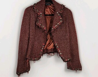 May include: A maroon tweed jacket with a herringbone pattern. The jacket has a frayed hem and is trimmed with gold-coloured embellishments. The interior lining is a solid rust colour. The jacket is hanging on a black hanger against a white background.