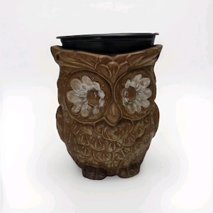 May include: A brown ceramic owl-shaped wax warmer featuring a sculpted feather texture and large, white-petaled flower designs around the eyes. The top of the owl figure is fitted with a removable black circular dish for holding wax melts.
