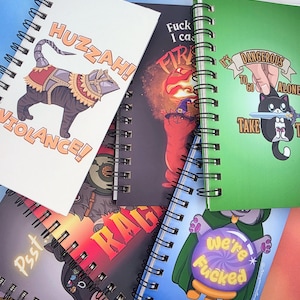 May include: A collection of spiral-bound notebooks featuring various cat-themed illustrations and text. Designs include a cat in armour with 'HUZZA! VIOLANCE!', a cat with a crystal ball saying 'we're fucked', and a cat holding a sword with 'It's dangerous to go alone'.
