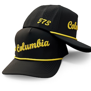 May include: Two black baseball caps with yellow embroidered text. One cap reads "Columbia" and the other reads "573" and "Columbia". Both caps have a yellow rope detail along the brim.