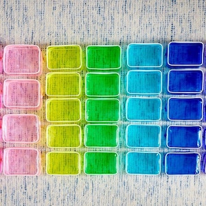 May include: A collection of rectangular, transparent containers arranged in a grid, showcasing a vibrant gradient of colors. The spectrum transitions from pink to green, then to blue and purple, with white borders.