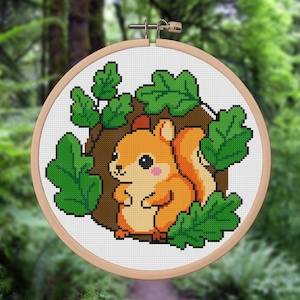 May include: A cross-stitch embroidery hoop featuring a cartoon squirrel. The squirrel is orange with a fluffy tail, pink cheeks, and black eyes. It is surrounded by green leaves and a brown tree trunk, all set against a white background.