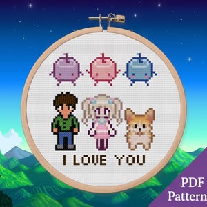 May include: A circular wooden embroidery hoop containing a cross-stitch design on white fabric. The pattern features three pixel-art style fruit characters, a male figure, a female figure, a dog, and the text I LOVE YOU in dark brown letters.