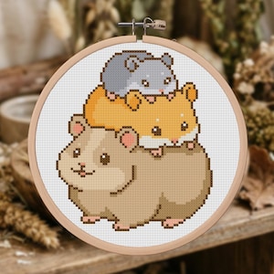 Op de afbeelding: Een ronde houten borduurring met een kruissteekpatroon van drie gestapelde hamsters. De bovenste hamster is grijs, de middelste is oranje en de onderste is lichtbruin. Elke hamster heeft kleine roze oortjes en donkere ogen op witte stof.