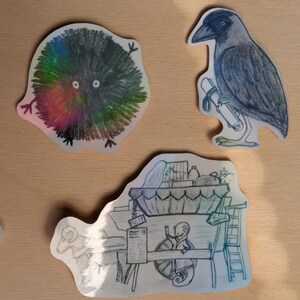 Könnte beinhalten: Eine Sammlung skurriler Aufkleber mit handgezeichneten Illustrationen. Enthält ein farbenfrohes, flauschiges Wesen, eine Krähe mit einer Schriftrolle und eine detaillierte Szene mit einem Wagen und Figuren. Die Aufkleber haben ein holografisches Finish.