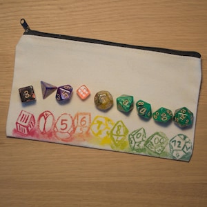 Könnte beinhalten: Eine weiße Canvas-Tasche mit Reißverschluss und einem Regenbogenmuster sowie einem Satz von 12 Würfeln. Die Würfel sind in verschiedenen Farben, darunter Grün, Lila und Gold.