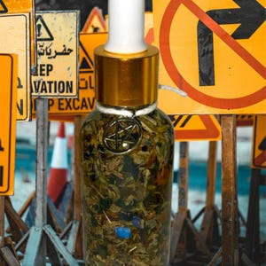 Könnte beinhalten: Eine Glasflasche mit goldenem Deckel und Tropfer. Die Flasche ist mit einer klaren Flüssigkeit und Kräutern gefüllt. Die Flasche steht vor einem verschwommenen Hintergrund aus gelben Baustellen Schildern.