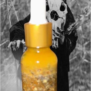 May include: A clear glass bottle with a gold cap and white dropper, filled with a yellow liquid containing visible particles. The background features a black and white image of a figure in a hooded cloak and a skull mask.