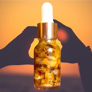 Puede incluir: Una botella de vidrio con tapa dorada y gotero llena de un l&iacute;quido amarillo y flores secas. La botella se sostiene entre dos manos contra un fondo de puesta de sol.