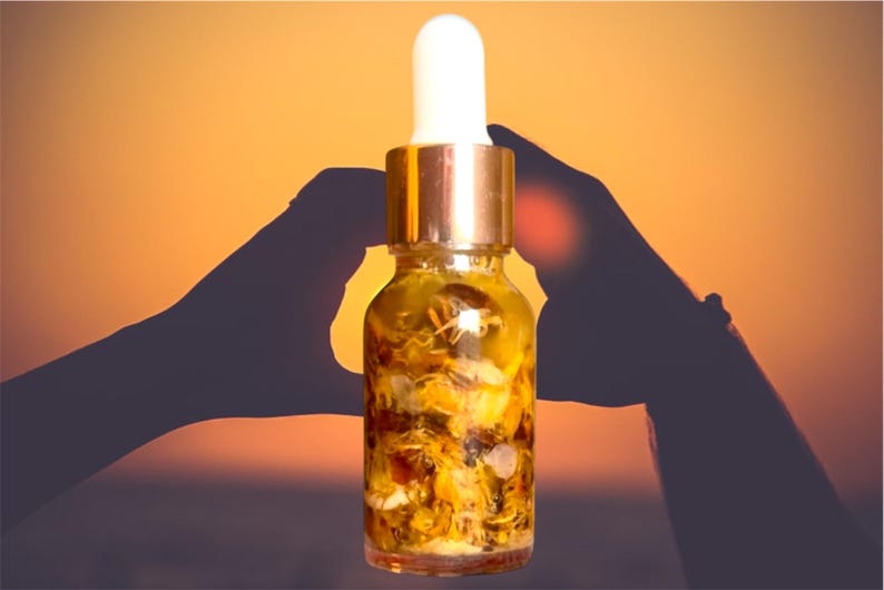 Puede incluir: Una botella de vidrio con tapa dorada y gotero llena de un l&iacute;quido amarillo y flores secas. La botella se sostiene entre dos manos contra un fondo de puesta de sol.