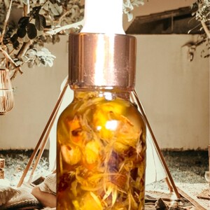 Puede incluir: Una botella de vidrio transparente con tapa dorada y gotero. La botella est&aacute; llena de un l&iacute;quido amarillo y contiene flores amarillas secas.