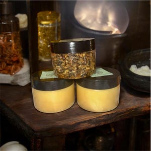 May include: Three jars of skincare products are stacked on a wooden shelf. Two jars contain a yellow cream, while the top jar holds a mixture of dried herbs. Additional jars with contents are visible in the background.