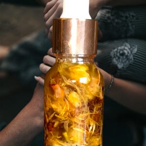 Puede incluir: Una botella de vidrio con tapa dorada y un gotero blanco llena de un l&iacute;quido amarillo y flores amarillas secas.
