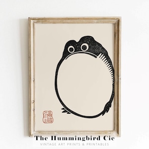 Peut inclure: Illustration en noir et blanc d'une grenouille dans un cercle, avec un tampon rouge dans le coin inférieur gauche. L'image est encadrée dans un cadre en bois clair. Le texte "The Hummingbird Cie" est sous le cadre.