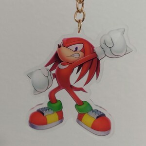 May include: A red and white cartoon character with spiky red hair and green shoes. The character is holding a fist in the air and has an angry expression on its face. The character is a keychain made of clear acrylic.