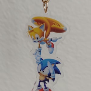 May include: A clear acrylic keychain featuring Sonic the Hedgehog and Tails. Sonic is blue with white gloves and red shoes. Tails is yellow with a white belly and red shoes. The keychain has a gold chain.