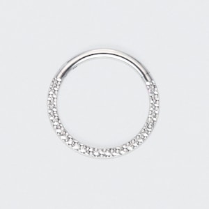 May include: A circular silver-toned hinged hoop ring featuring a textured surface with numerous small, reflective facets that create a shimmering effect. The jewelry piece has a seamless closure mechanism visible at the top of the circular frame.