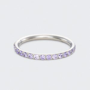 May include: A silver ring featuring a continuous line of small, light purple gemstones. The ring is circular and set against a white backdrop. The gemstones are uniformly spaced around the band.