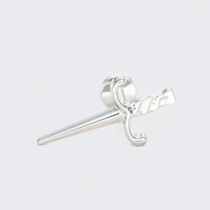 Op de afbeelding: Een zilverkleurige reversspeld in de vorm van een klein zwaard. De speld heeft een gedetailleerde handgreep en een puntig lemmet. De speld is ontworpen om op kleding te worden gedragen, zoals een jas of blazer.