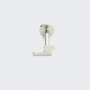 May include: A silver-colored lip piercing with a flat, geometric design. The piercing has a flat disc on top, a straight post, and a stylized lightning bolt shape at the bottom. The jewelry is set against a white background.