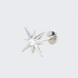 May include: A silver ear piercing stud with a starburst design. The starburst has multiple pointed rays and a small, clear gemstone in the centre. The stud has a flat, round backing.