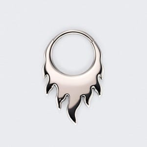 May include: A silver-colored, flame-shaped body jewelry piece. The jewelry has a circular top with a hinged closure and a stylized flame design extending downwards. The metal has a polished, reflective surface.