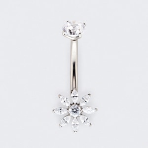 Può includere: Un piercing all'ombelico argentato con una grande gemma trasparente e sfaccettata nella parte superiore e un motivo floreale nella parte inferiore. Il fiore è composto da più pietre chiare a forma di marquise che circondano una pietra centrale rotonda.