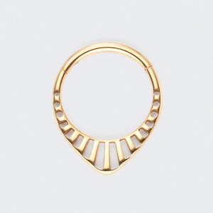 Puede incluir: Un anillo de tabique dorado con un diseño decorativo en forma de abanico. El anillo tiene forma circular con una serie de pequeños agujeros y barras verticales en la parte inferior, creando un patrón geométrico ornamentado. La joya está sobre un fondo blanco.