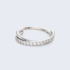 May include: A silver ring with a double-band design. The lower band is set with small, clear gemstones. The ring is displayed against a plain white background, emphasizing its polished metal and sparkling stones.
