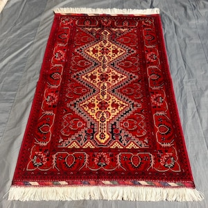 Puede incluir: Una alfombra rectangular anudada a mano con un campo rojo intenso y intrincados patrones geométricos en crema, negro y dorado. La alfombra presenta un diseño de medallón central y una franja blanca en los extremos cortos. Esta alfombra es adecuada para la decoración del hogar.