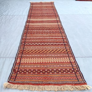 Puede incluir: Una alfombra pasillera larga y rectangular con un patrón repetitivo de rayas horizontales en tonos rojo intenso, naranja y crema. La alfombra está decorada con intrincados motivos geométricos, incluyendo diamantes y zigzags, y rematada con flecos color crema.