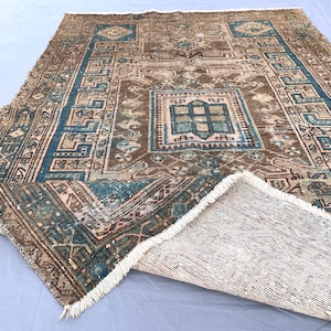 May include: A rectangular area rug featuring a central geometric medallion in shades of brown, teal, and cream. The border displays a repeating Greek key pattern in teal and brown. One corner is folded over, revealing the woven backing and white fringe.