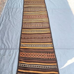 Puede incluir: Una alfombra de pasillo larga y rectangular con un patrón tejido de rayas horizontales en tonos marrón, beige y amarillo dorado. El diseño incluye motivos geométricos de diamantes y pequeños acentos cuadrados, con un borde marrón oscuro y flecos.