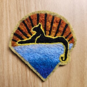 May include: A needle felted patch featuring a black silhouette of a reclining panther centered against a vibrant orange and yellow sunburst pattern, positioned above a textured blue landscape, all bordered by a light yellow felted edge.