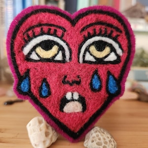 May include: A needle-felted heart-shaped patch in vibrant magenta with a black outline. The design features two large eyes with yellow irises, black pupils, and long lashes, two blue teardrops, a small nose, and an open mouth showing two white teeth.