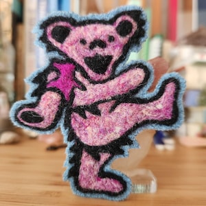 May include: A fuzzy, embroidered patch shaped like a dancing bear. The bear features a mottled pink and magenta body with thick black outlines, black ears, a black nose, and a black mouth. The entire figure is bordered by a light blue edge.