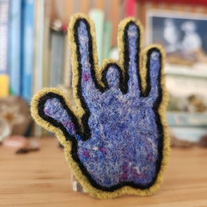May include: A needle-felted hand gesture patch in blue with purple and white flecks. The hand shape is outlined in a thick black border and finished with a mustard yellow edge. The design depicts the American Sign Language sign for I love you.