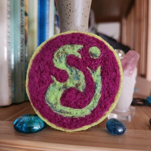May include: A round, felt pin with a green and yellow design on a deep pink background. The design is a stylized swirl.