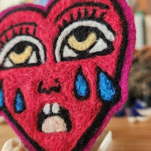 May include: A heart-shaped needle felted patch in vibrant red with a dark pink border. The design features two large eyes with black lashes, yellow irises, and white sclera, a small nose, a mouth with two white teeth, and three blue teardrops.